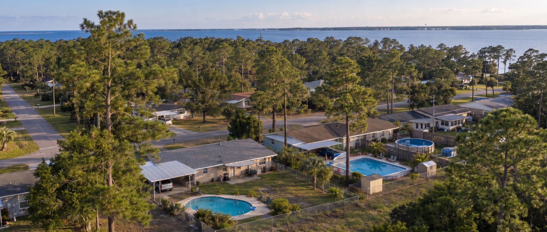 A low aerial over Callaway, with backyard pools behind single story ranch homes and carports in the foreground under heavy pine cover