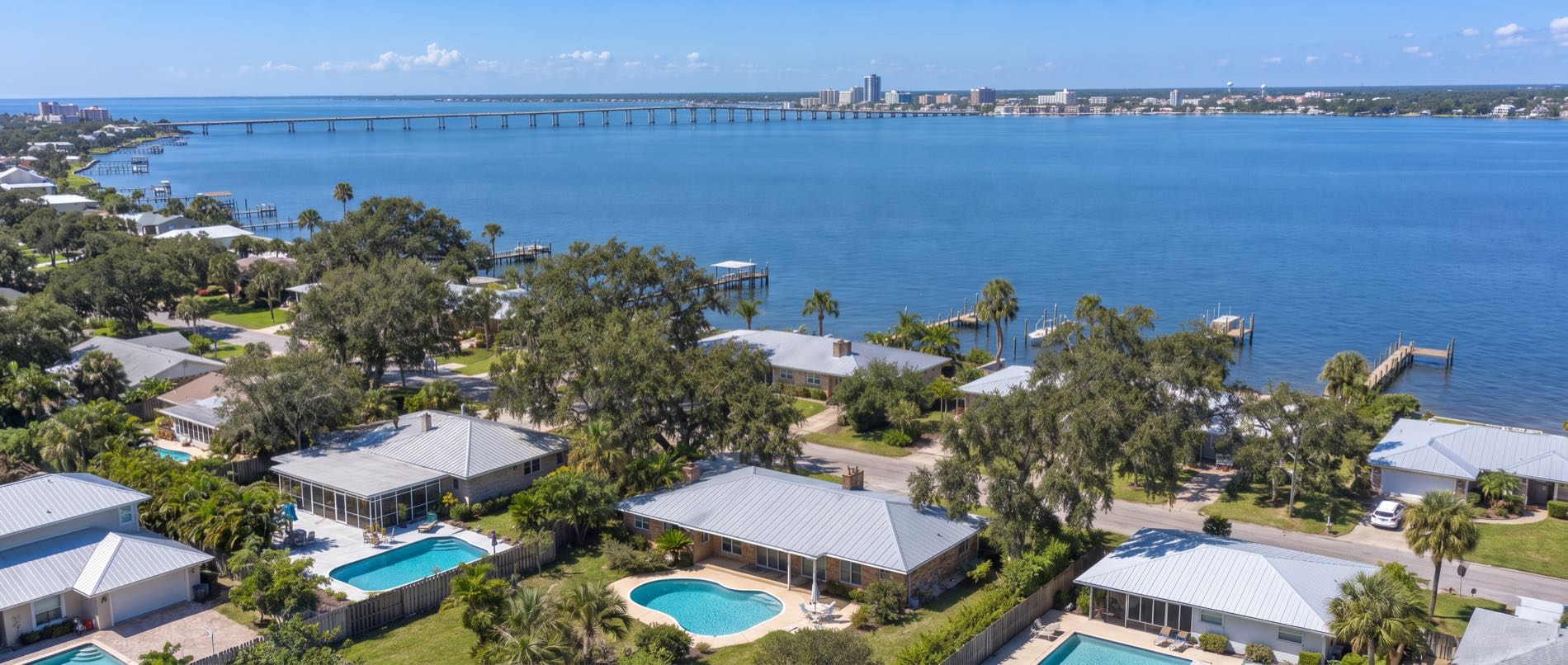 A low aerial over Panama City, with backyard pools behind brick ranch homes in the foreground and live oaks running down to St Andrews Bay