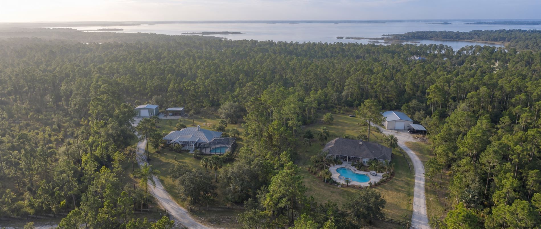 A low aerial over Southport, with pools on wooded acre lots in the foreground, long driveways through the pines and North Bay in the distance