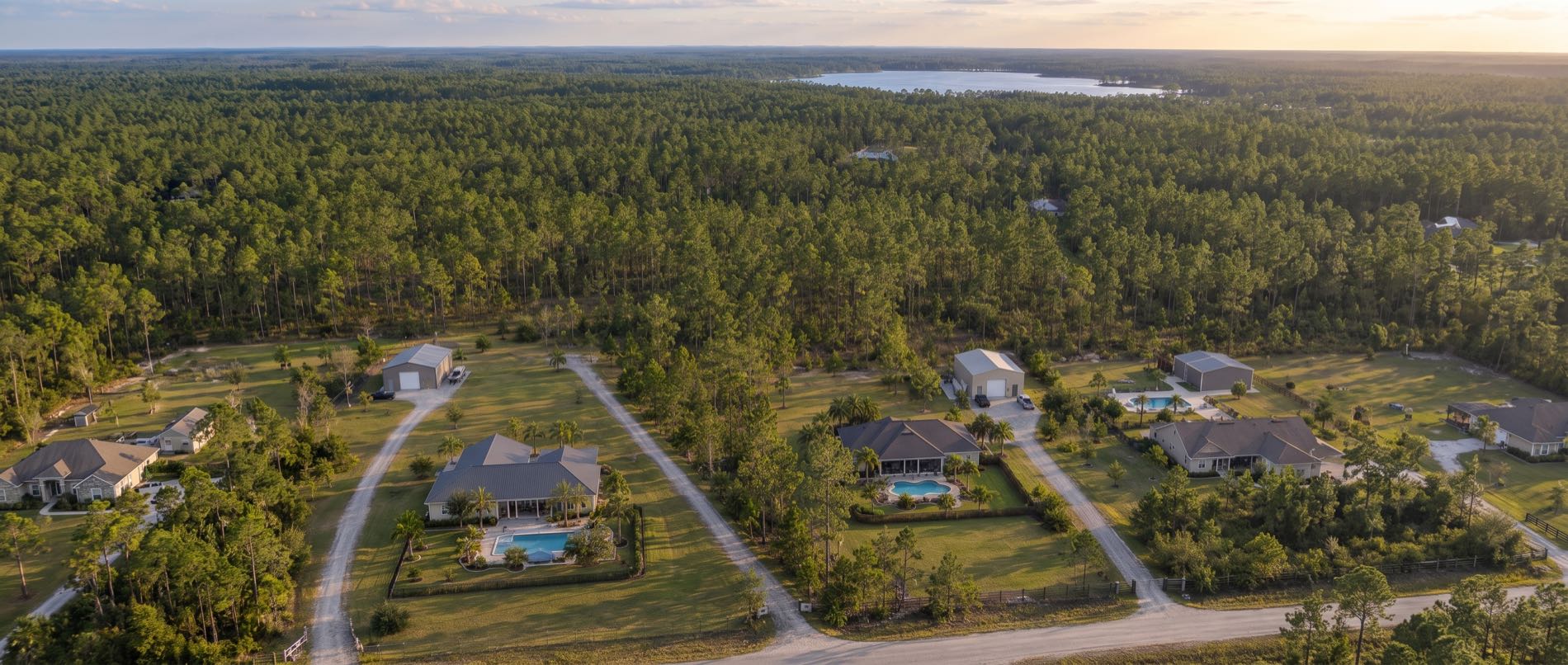 A low aerial over Youngstown, with a pool on a big cleared country lot in the foreground, a metal workshop beside the house and pine forest beyond