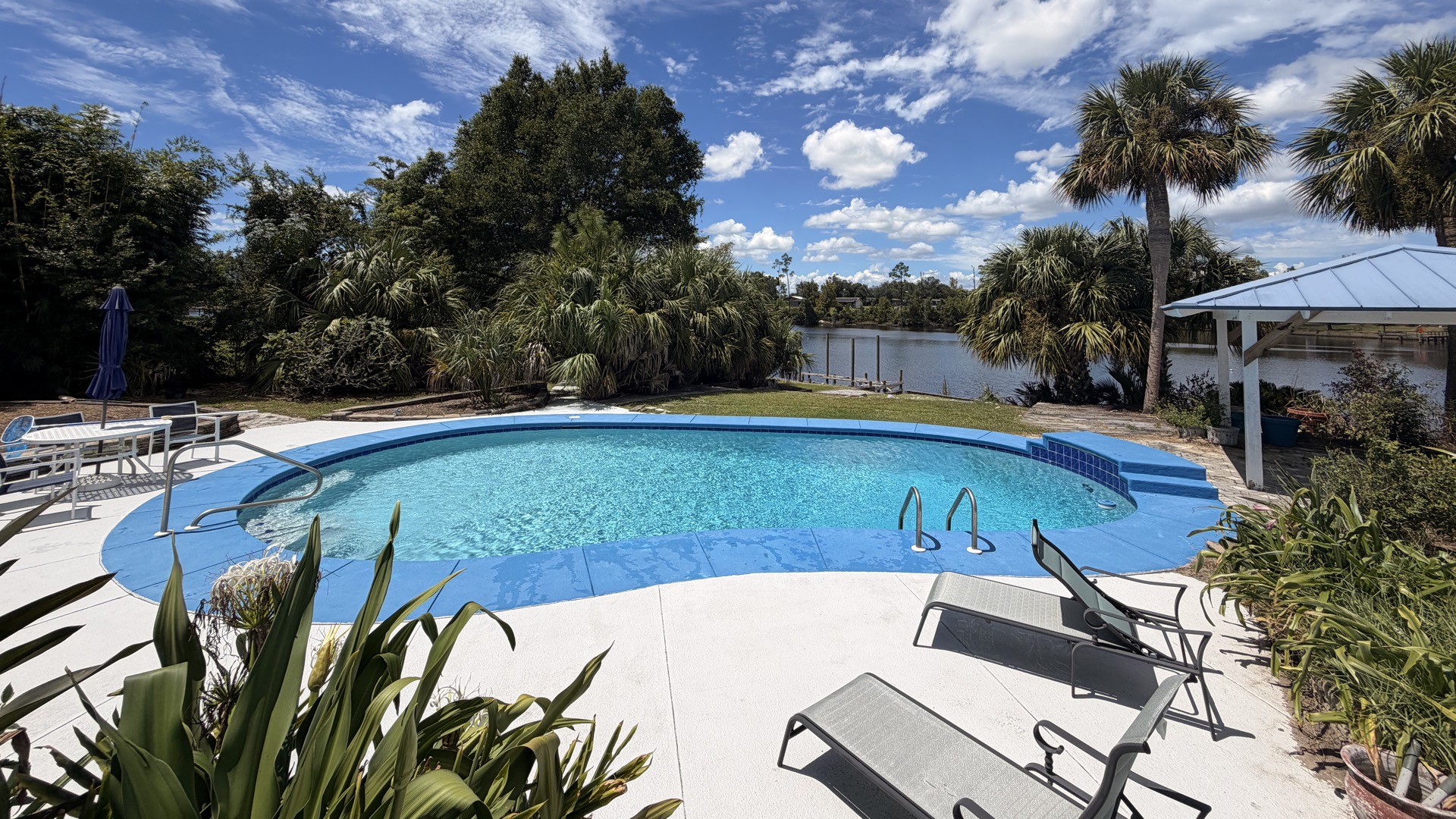 A clear blue kidney shaped pool on the water in Bay County, Florida, with palms, a covered dock and open sky behind it