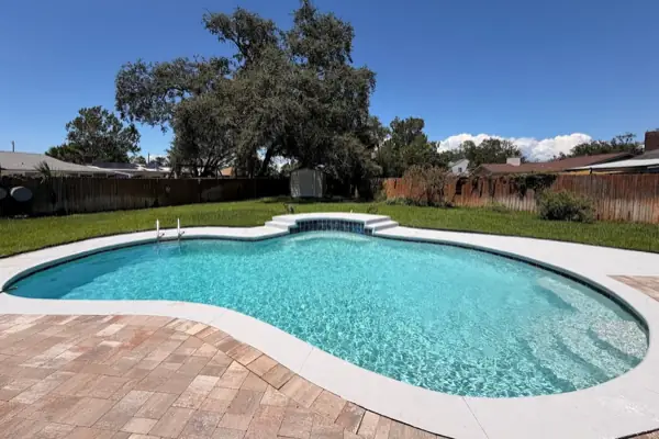 A clear kidney shaped pool with white coping and a blue tile step, under a big live oak in a fenced backyard