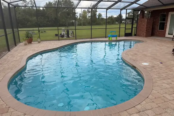 A clear kidney shaped pool with a paver deck inside a screen enclosure, looking out over a field