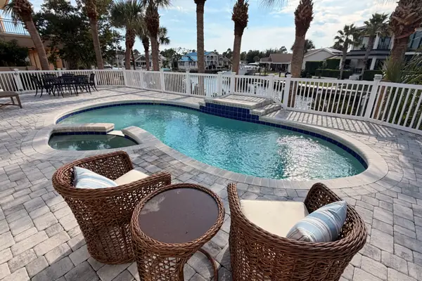 A kidney shaped pool and attached spa on a paver deck, with wicker chairs in front and palms and waterfront homes behind a white fence